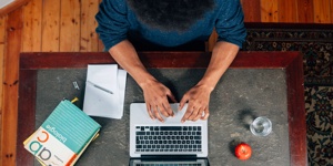 Eine Person sitzt an einem Schreibtisch und schreibt an einem Laptop. Auf dem Schreibtisch liegen unter anderem Notizzettel und Stifte.